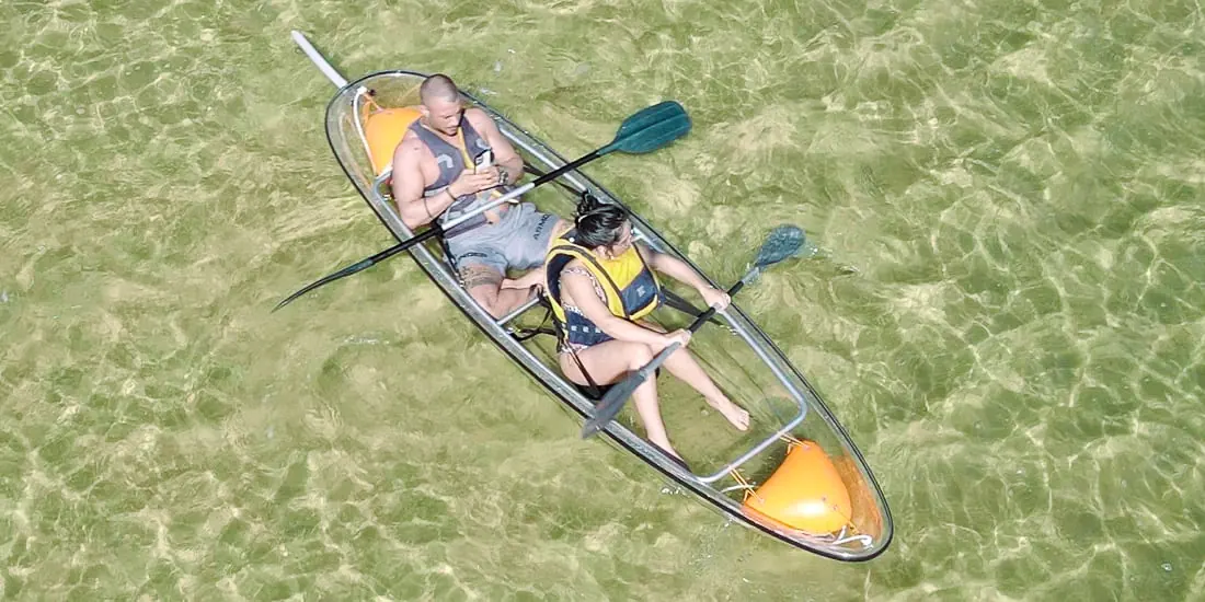 Two people seen from above in a transparent double kayak, the sandy lagoon floor clearly visible through the hull