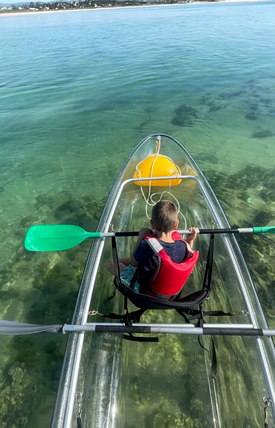 View through the clear hull of a kayak onto seagrass and sand, a child paddling