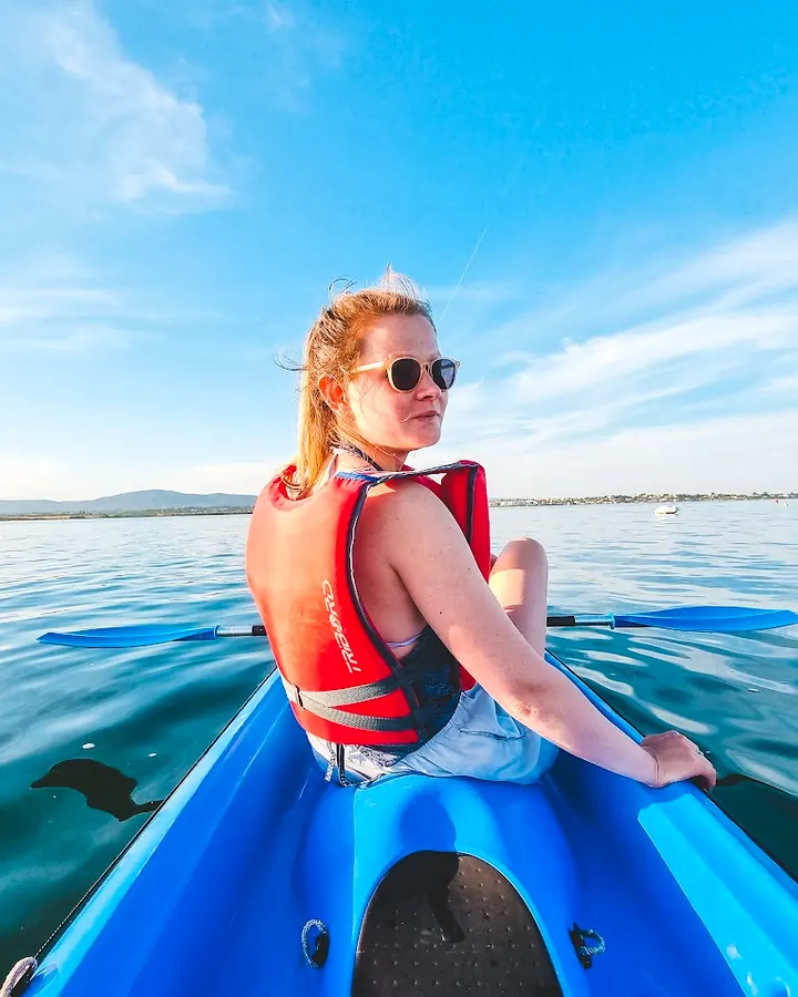 A woman in a red life jacket sitting in a blue kayak, looking back over calm water