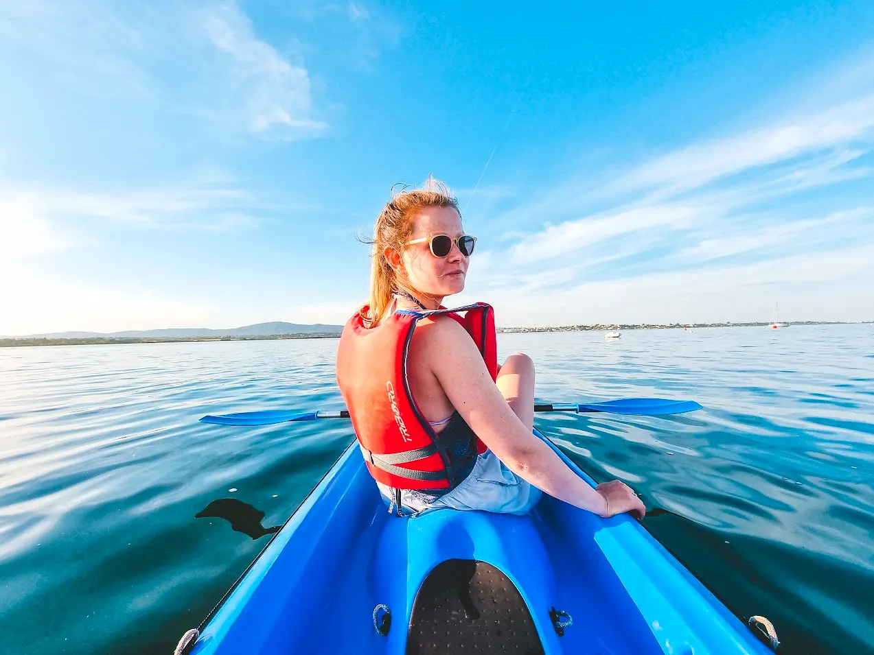 A paddler in a red life jacket sitting in a blue kayak, looking back over calm lagoon water